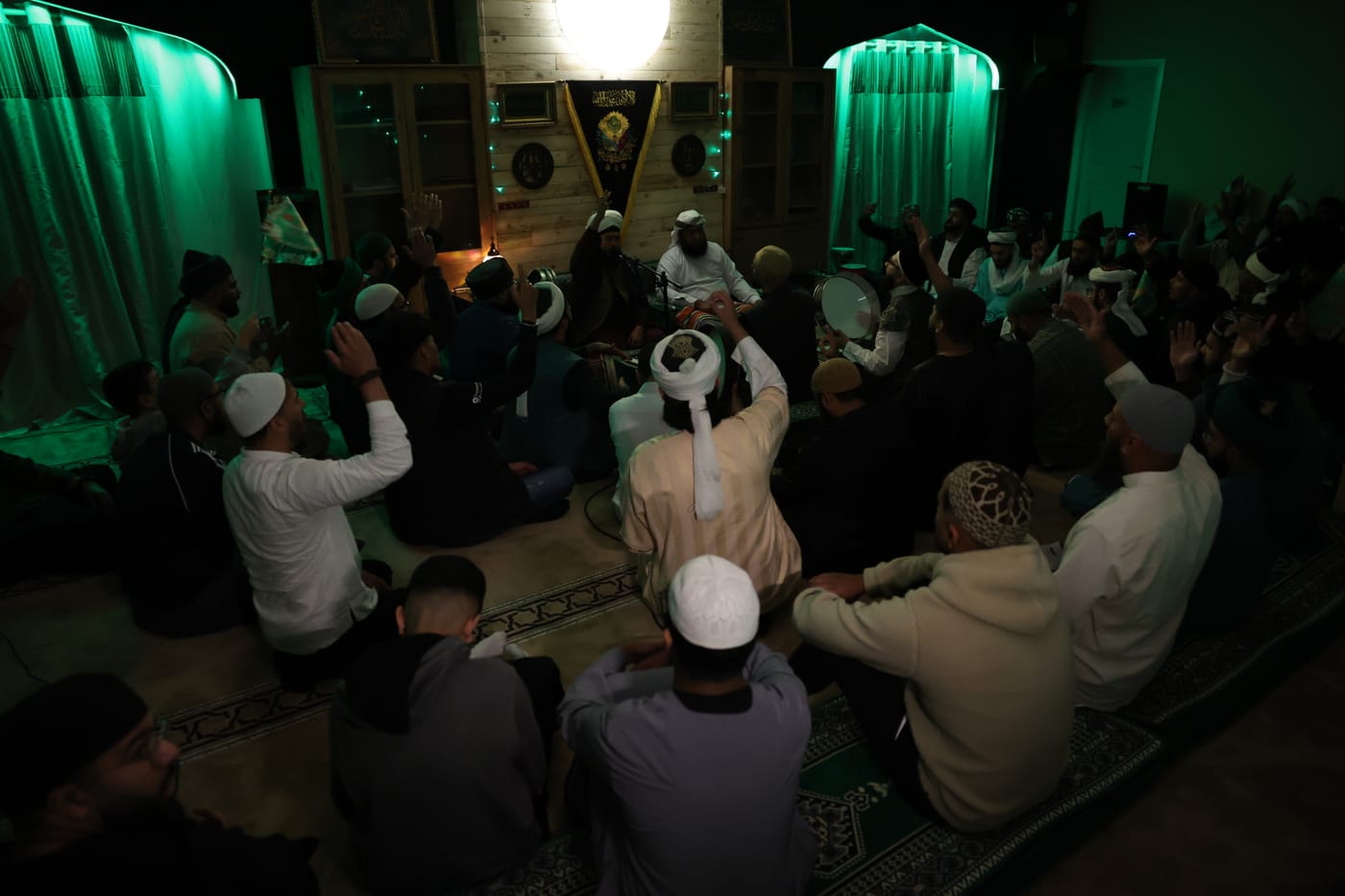 Murids raising their hands in dhikr under green light.
