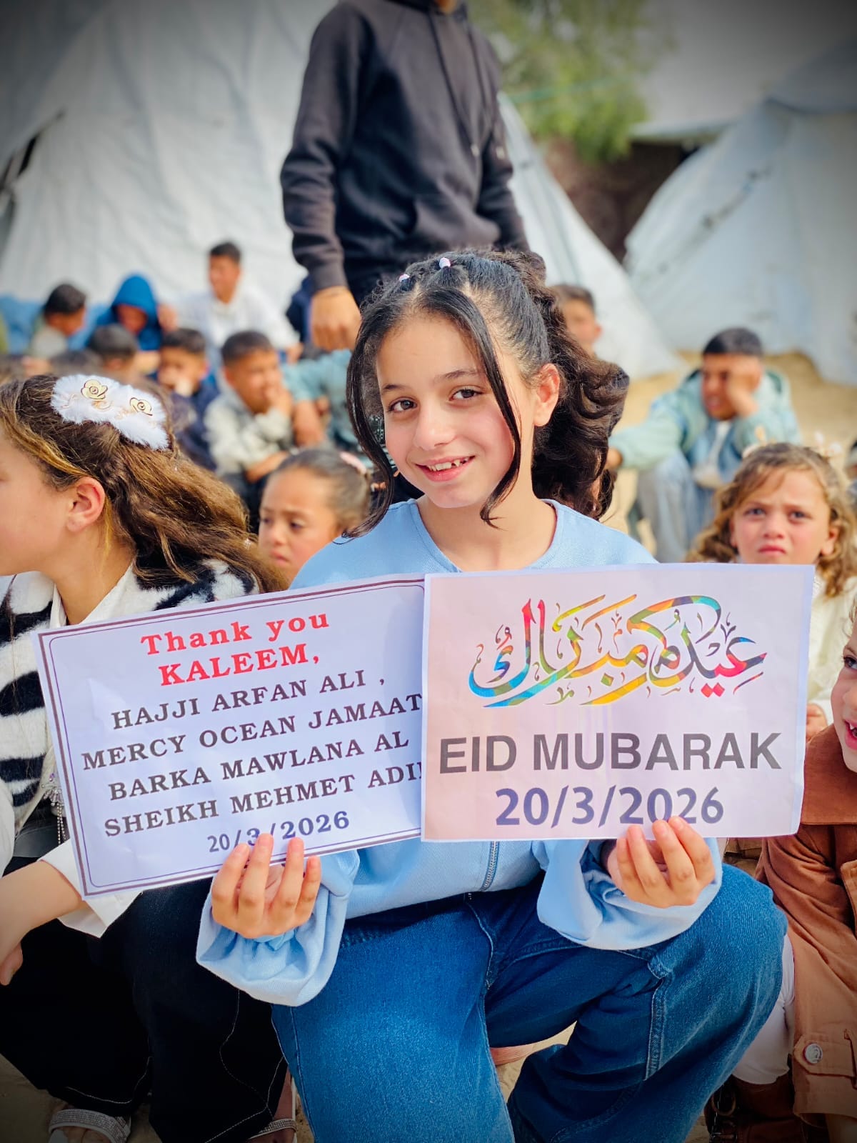 A girl in Gaza at Eid 2026, holding an Eid Mubarak sign and a sign thanking Mercy Ocean Jamaat and Maulana Shaykh Mehmet Adil.